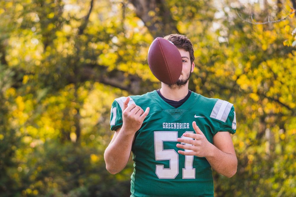 Football player in green jersey number 51 tosses ball upward against autumn foliage background.