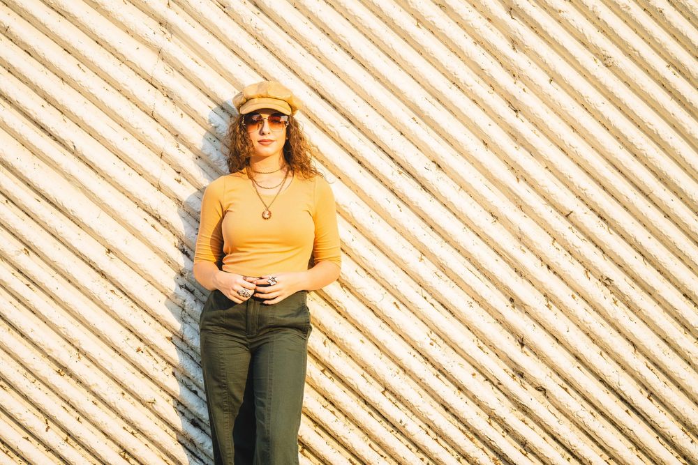 Someone in sunglasses and sun hat poses against a striped wall in golden sunlight wearing a yellow top and olive pants.