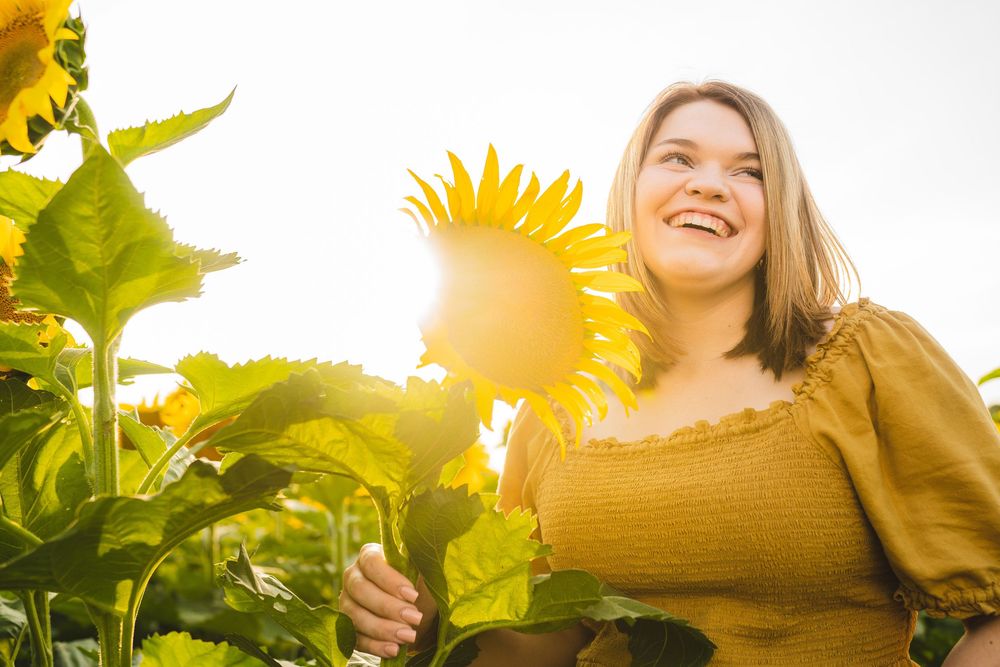 A person stands joyfully in a sunflower field at sunset, wearing a warm brown top against bright yellow blooms.
