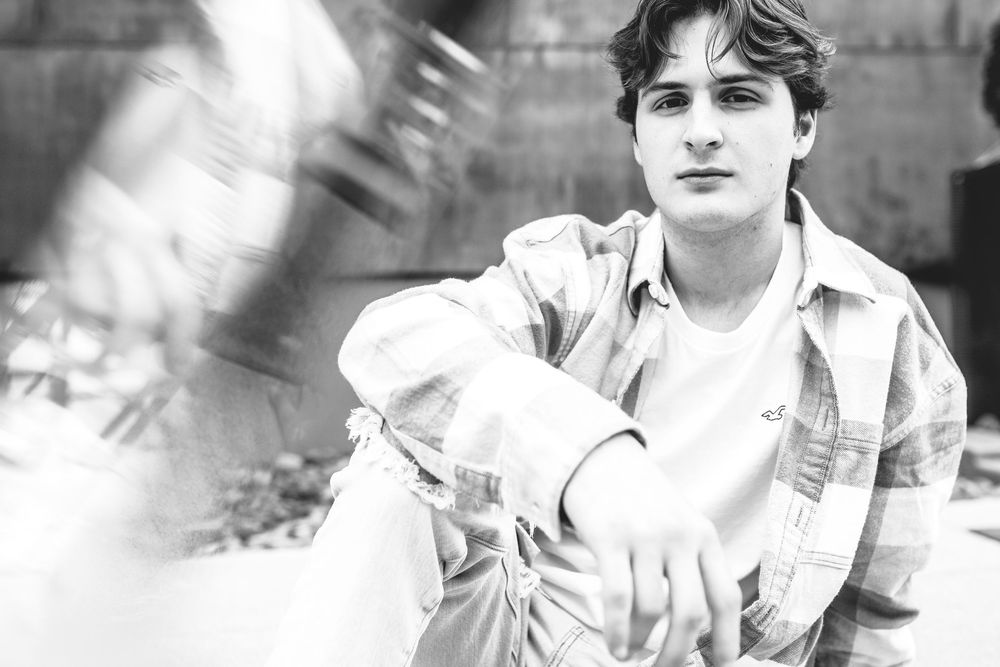 A black and white portrait photograph showing someone in a casual white shirt leaning against a wall.