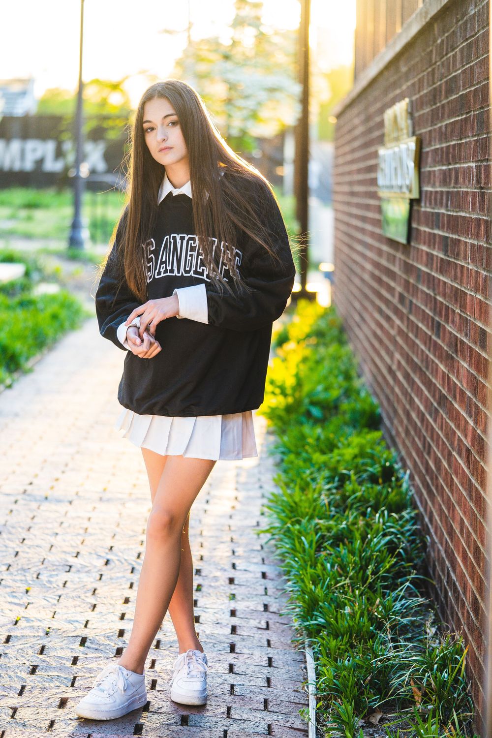 A person in black attire and white sneakers poses along a brick wall walkway with lush greenery.