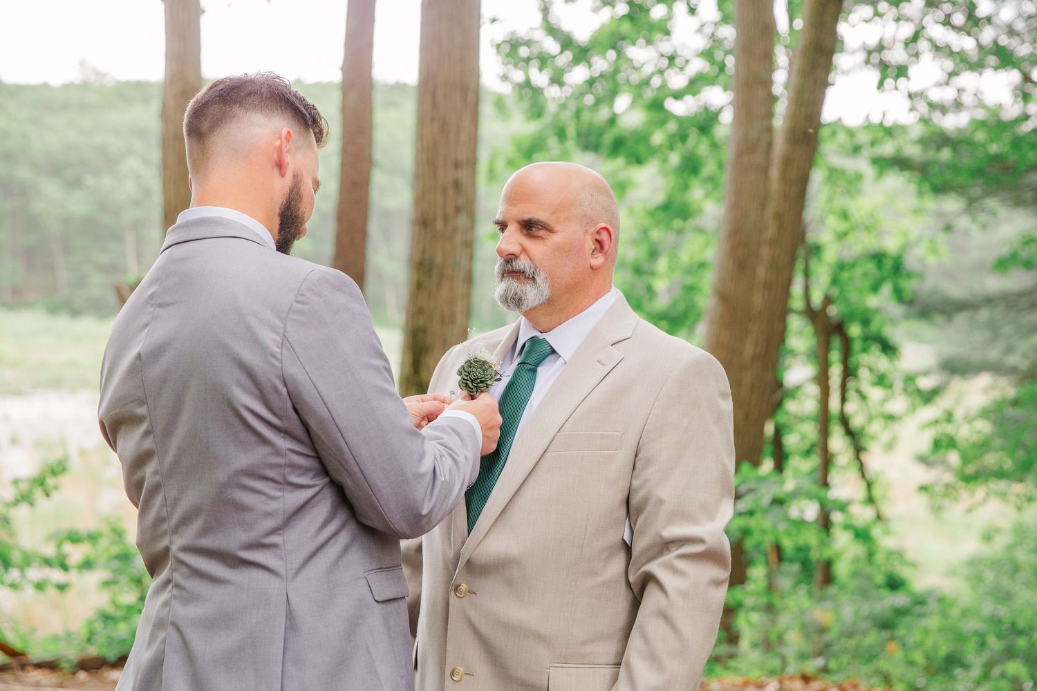 Three people in formal attire pose together with floral bouquets against a sunlit forest backdrop at a wedding celebration.