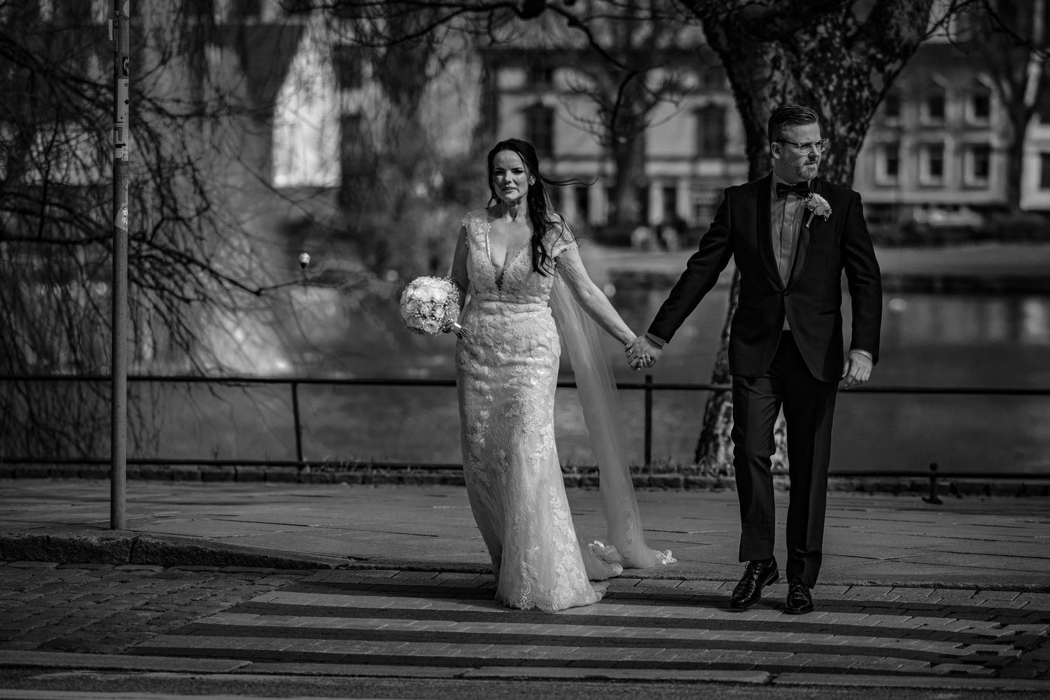 Black and white photo of elegant bride and groom walking hand in hand along riverside promenade.