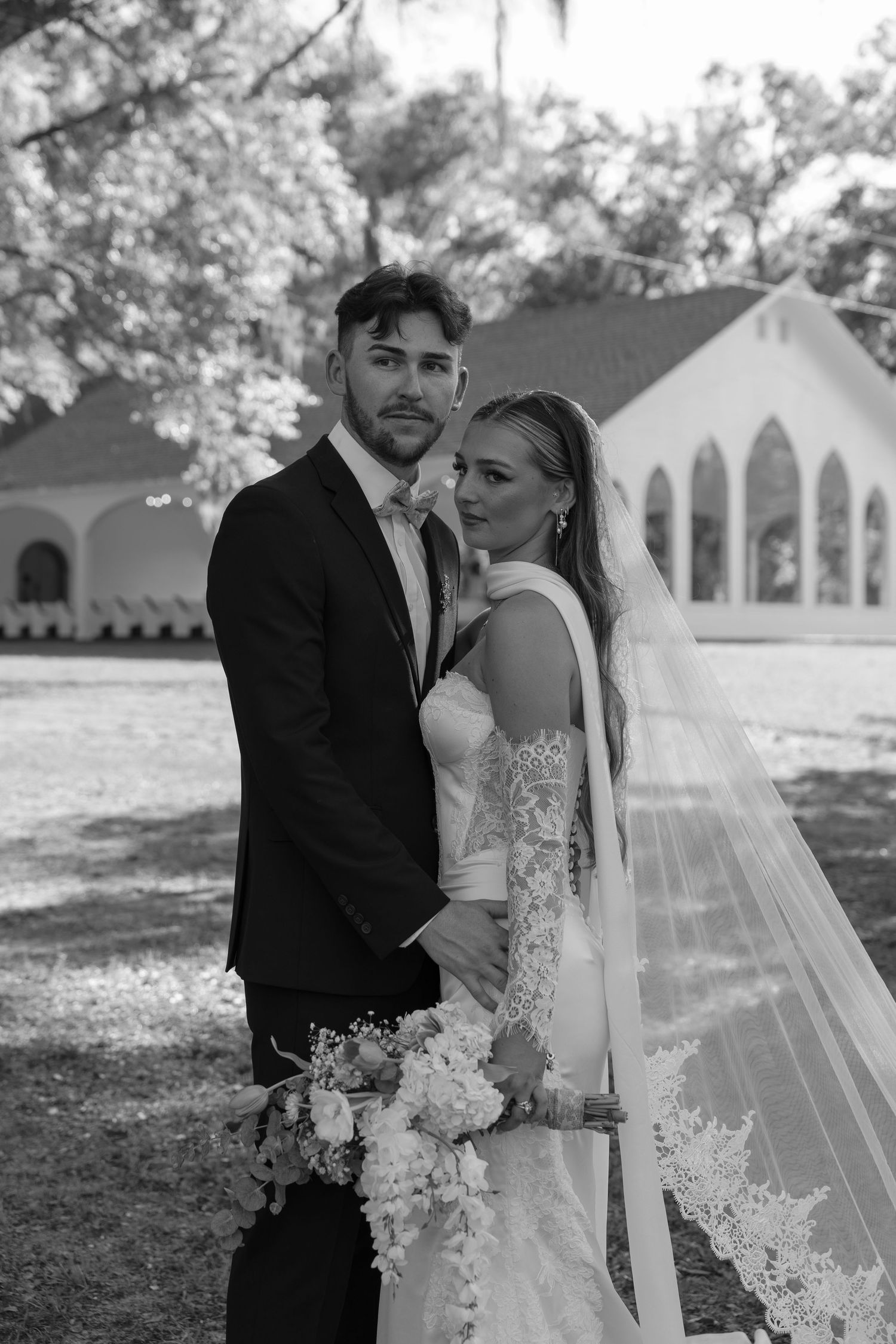 Two brides in white wedding gowns embrace romantically in a lush green garden surrounded by manicured hedges.
