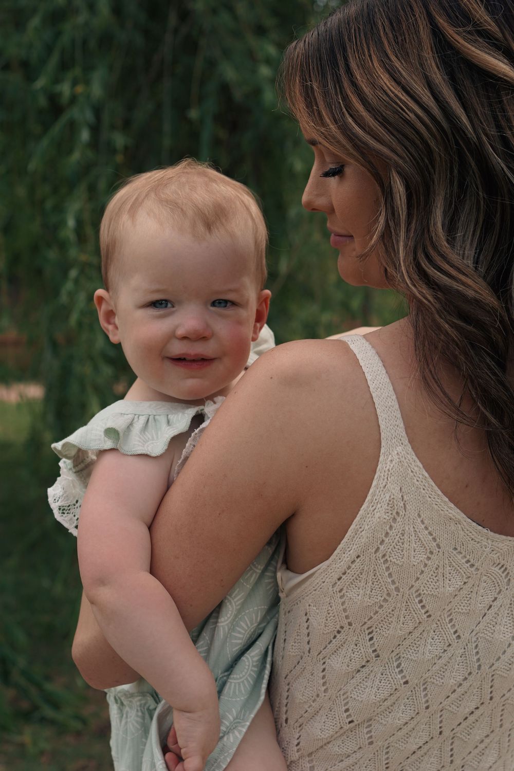 A tender moment captured outdoors as a family member holds a smiling baby wearing a ruffled outfit against a green background.