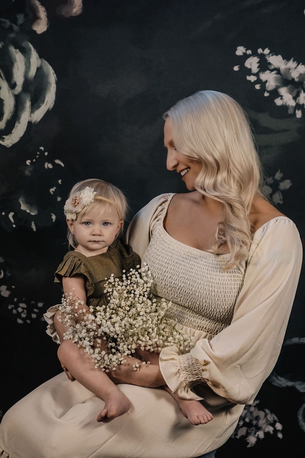 A touching family photoshoot with soft lighting features delicate baby's breath flowers and cream colored outfits against a dark backdrop.
