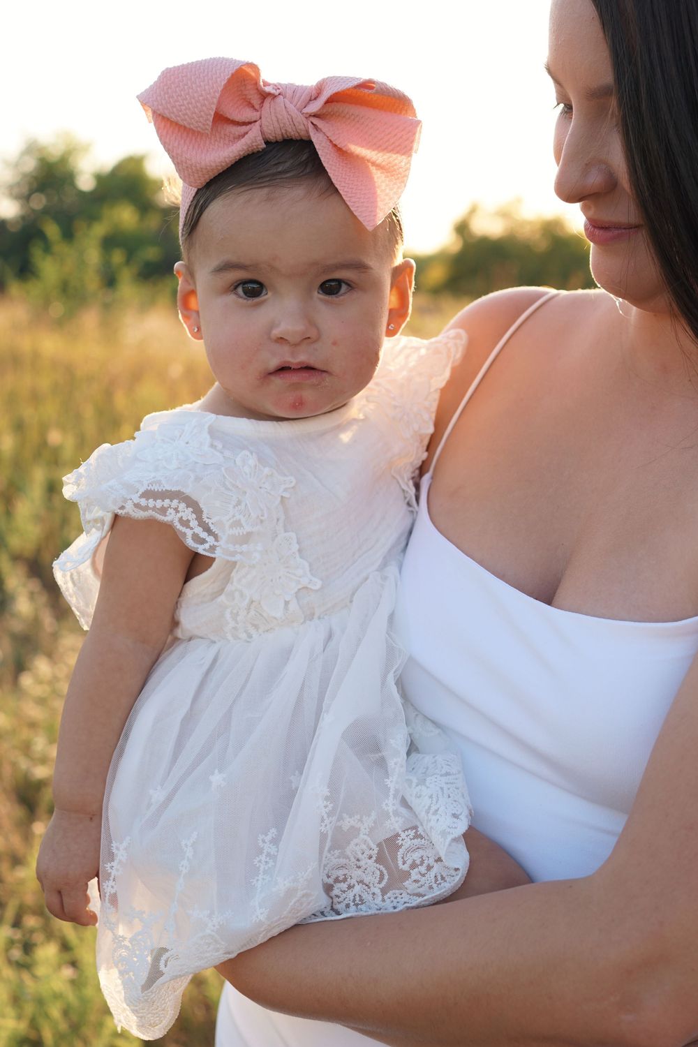 A baby wearing a white lace dress and pink bow headband is held outdoors during golden hour.