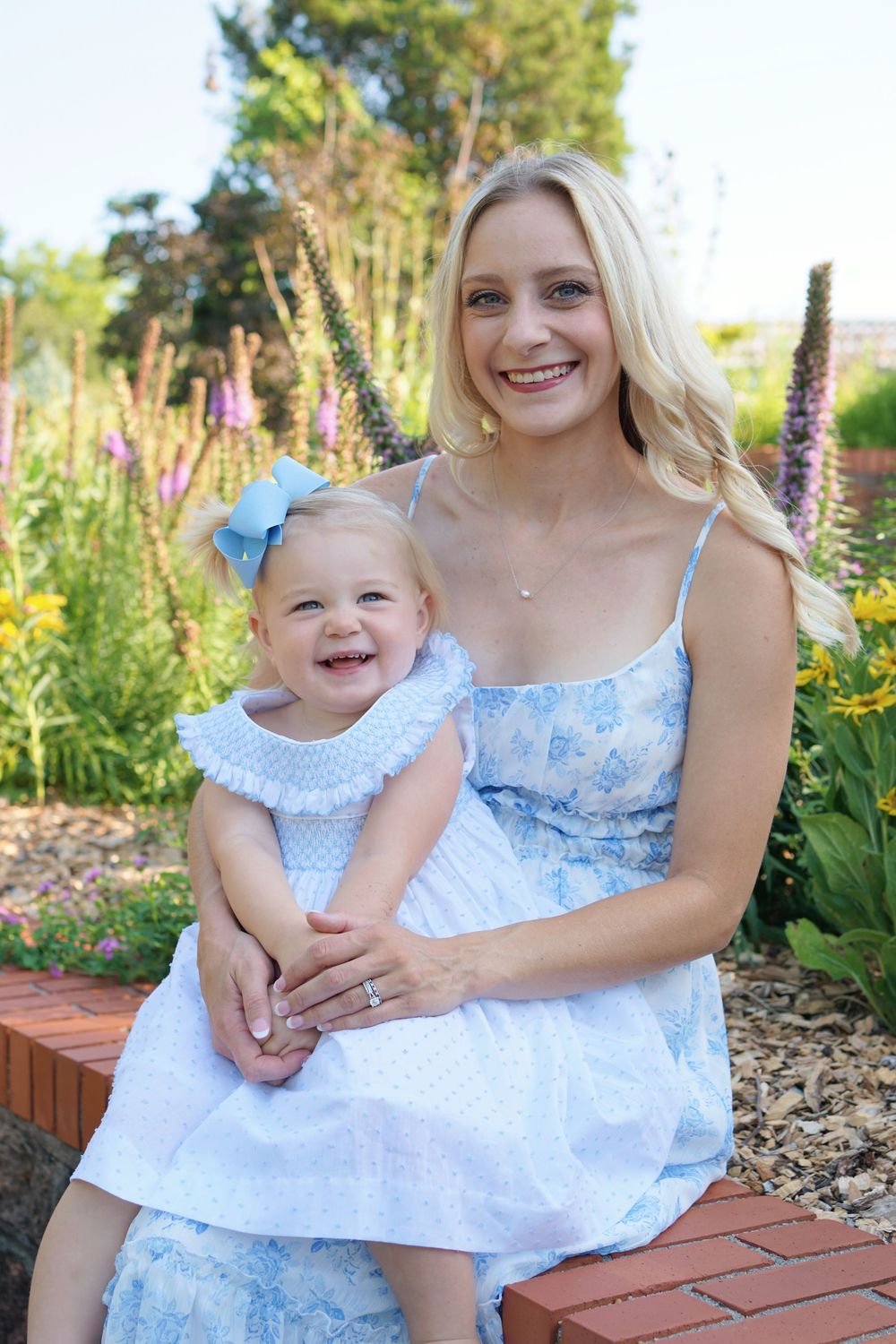 Mother and baby in matching blue and white dresses pose together on brick ledge in a garden setting.