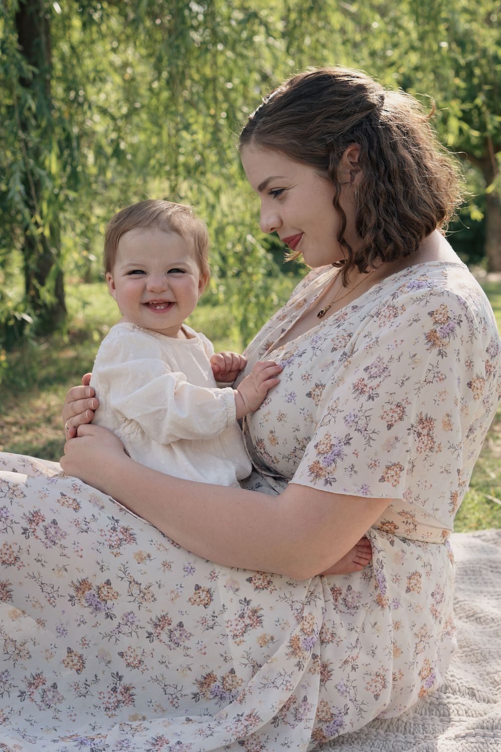 Mother and baby in white dresses share tender moment together while sitting outdoors beneath a willow tree.