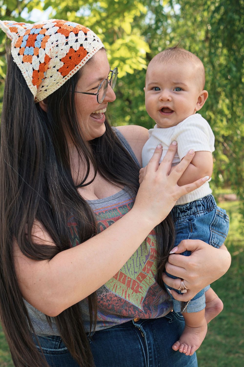 A parent wearing glasses and a colorful headscarf shares a joyful moment outdoors with their baby in summer.