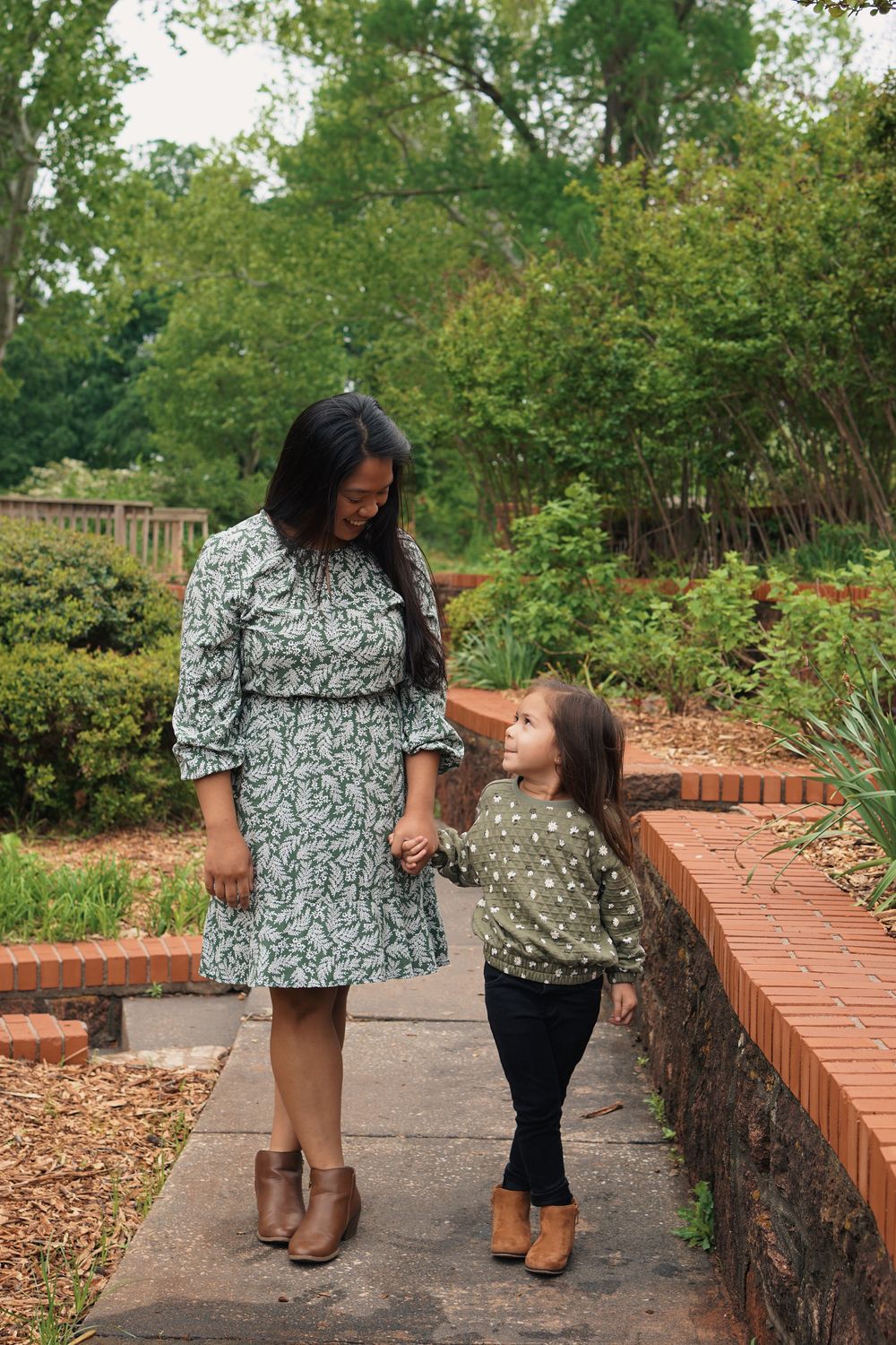 A parent and child wearing matching floral outfits stand together on a brick path in a lush garden setting.