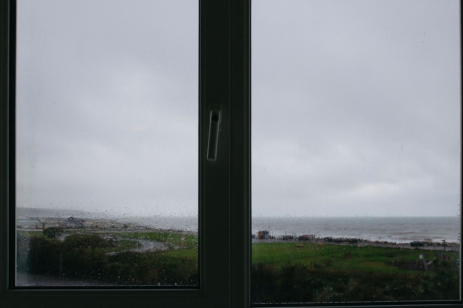 Cloudy coastal landscape viewed through a dark window frame with rolling green hills and ocean in the distance.