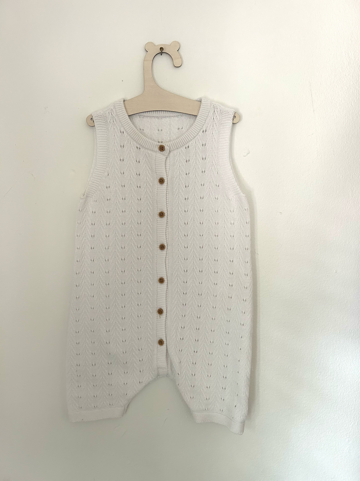 Vintage-style beige knitted baby romper with button details and side pockets hanging on display.