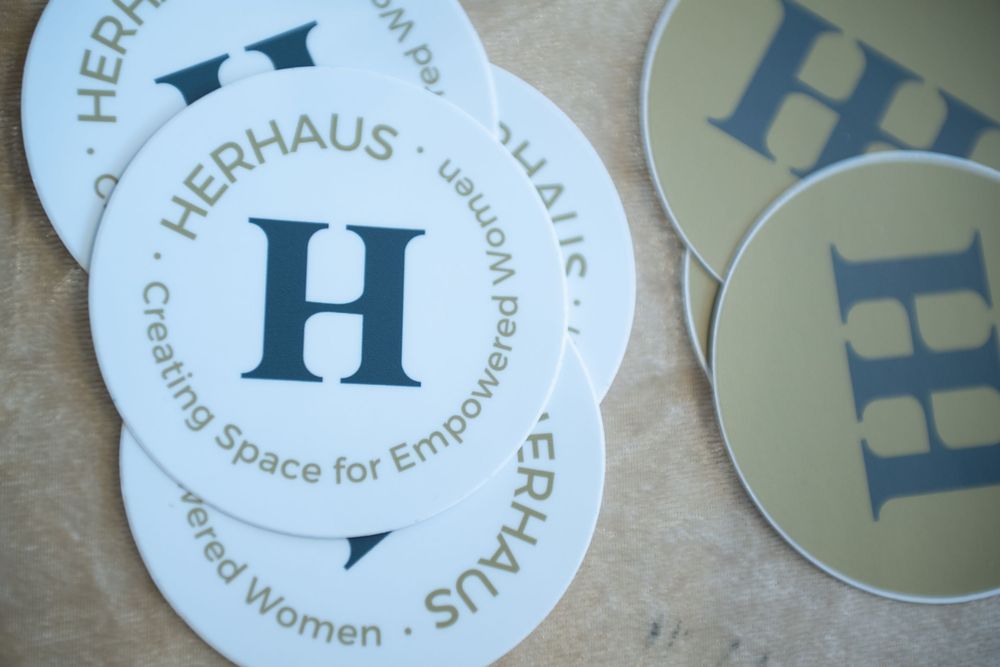White and navy blue circular Herrhaus coasters with H logo and text 'Creating Space for Empowered Women' displayed.