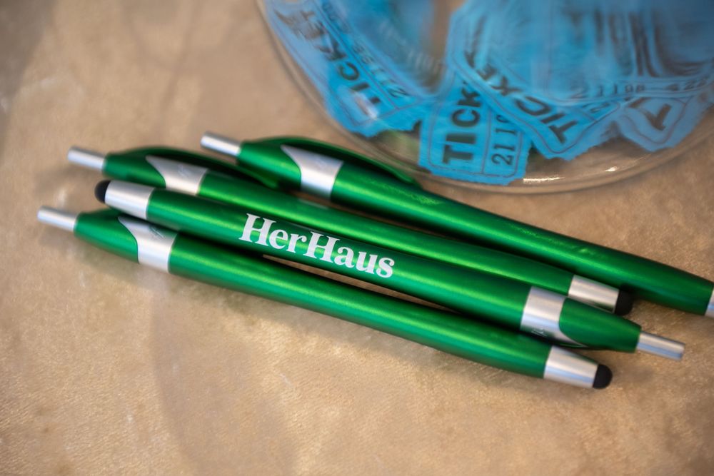 Three green metallic pens with 'HerHour' branding and silver accents sit on a wooden surface beside a blue object.