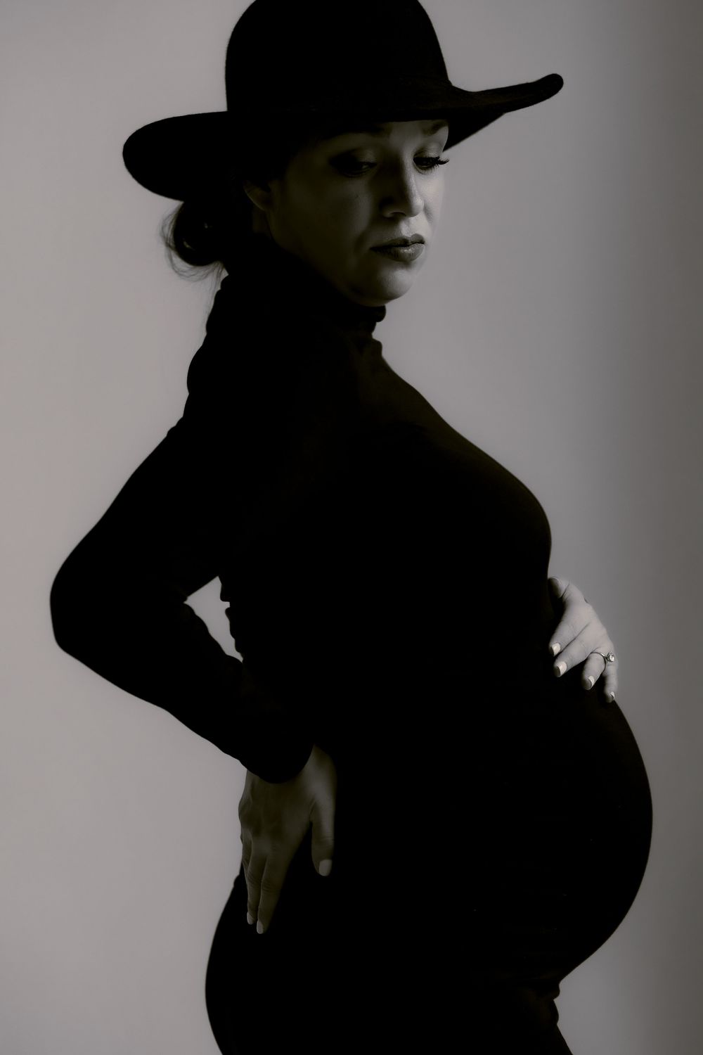 Silhouette of pregnant woman in black turtleneck and wide-brimmed hat poses against light background in artistic portrait.