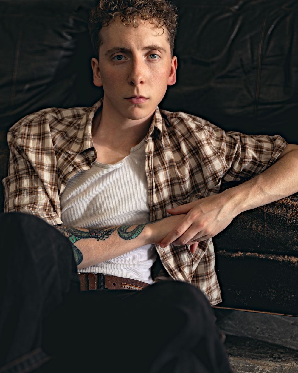 A young person in a plaid button-down shirt over white tank top leans against a dark background, revealing tattoos.