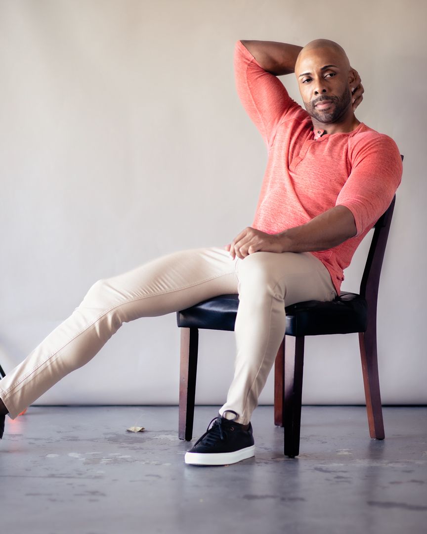 Stylish person in coral sweater and beige pants poses casually on wooden chair against neutral wall.