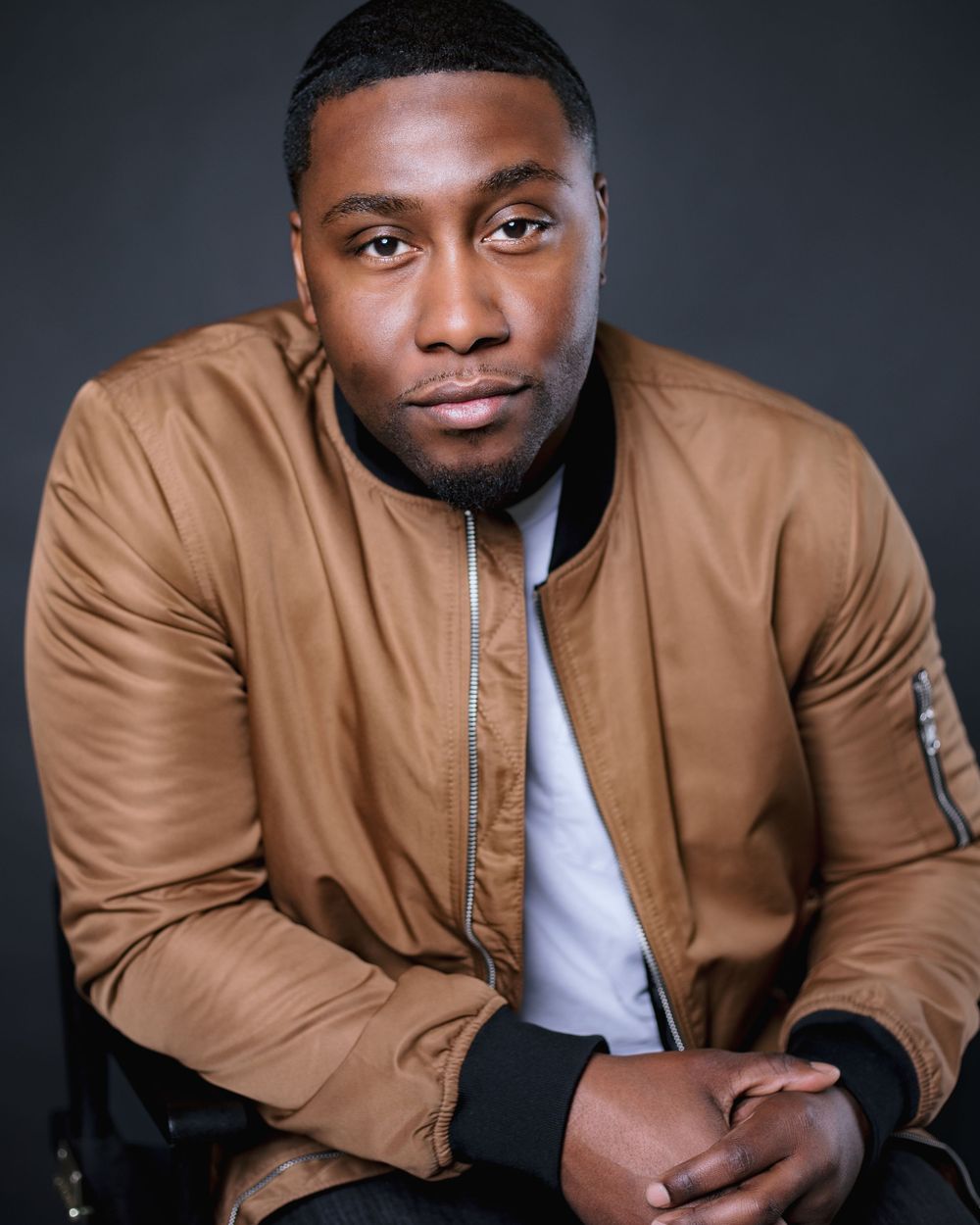 Professional headshot of someone wearing a tan bomber jacket over a white shirt against a dark background.