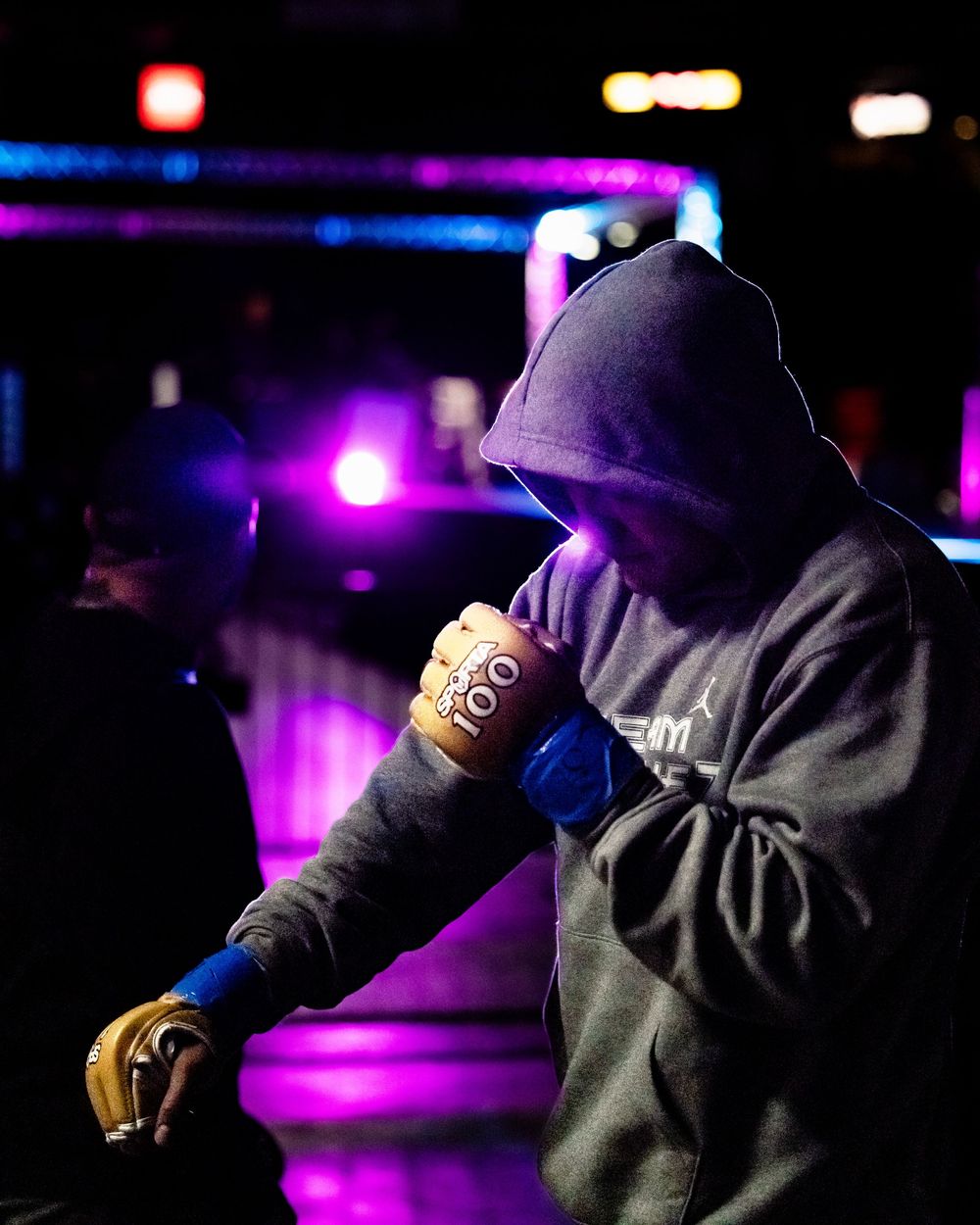 Figure in dark hoodie performs against vibrant purple and blue stage lighting at a nightclub venue.