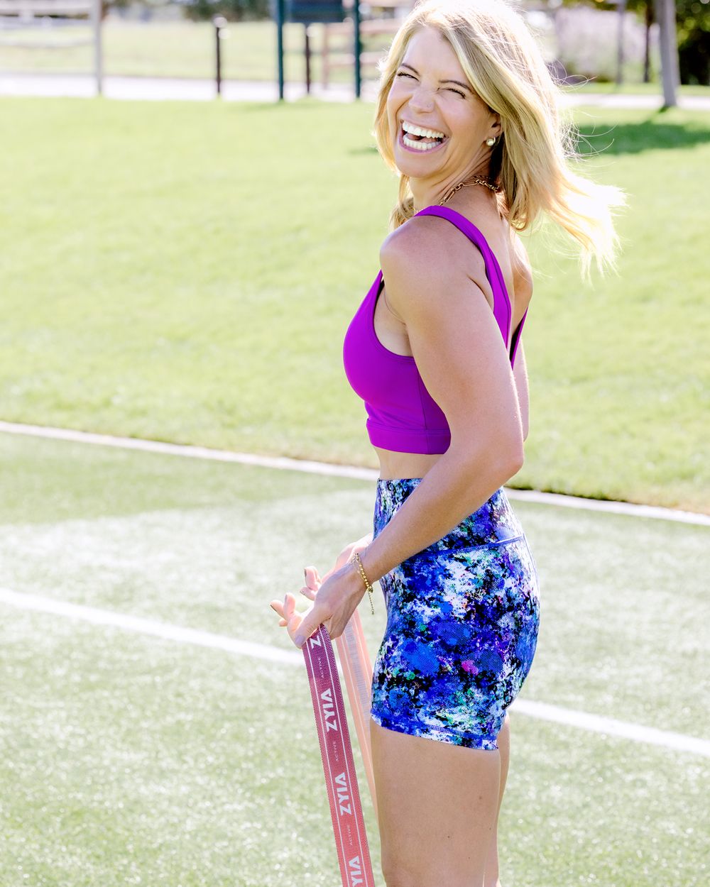 Athletic figure in purple tank top and floral blue shorts stands on sports field with resistance band for exercise.