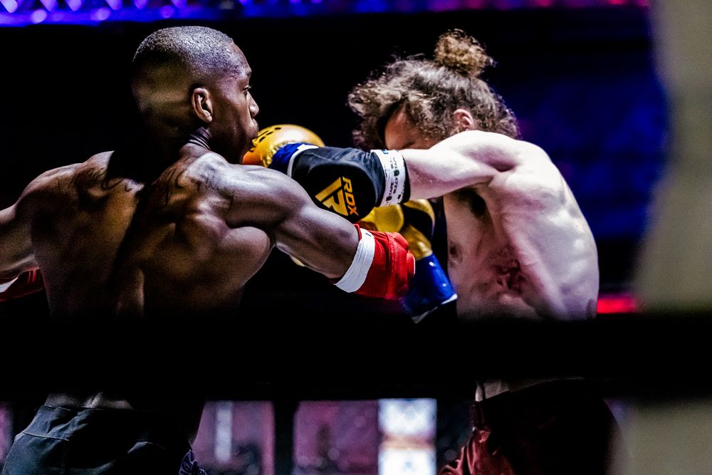 Two boxers exchange punches during an intense boxing match in a dark arena with purple lighting.