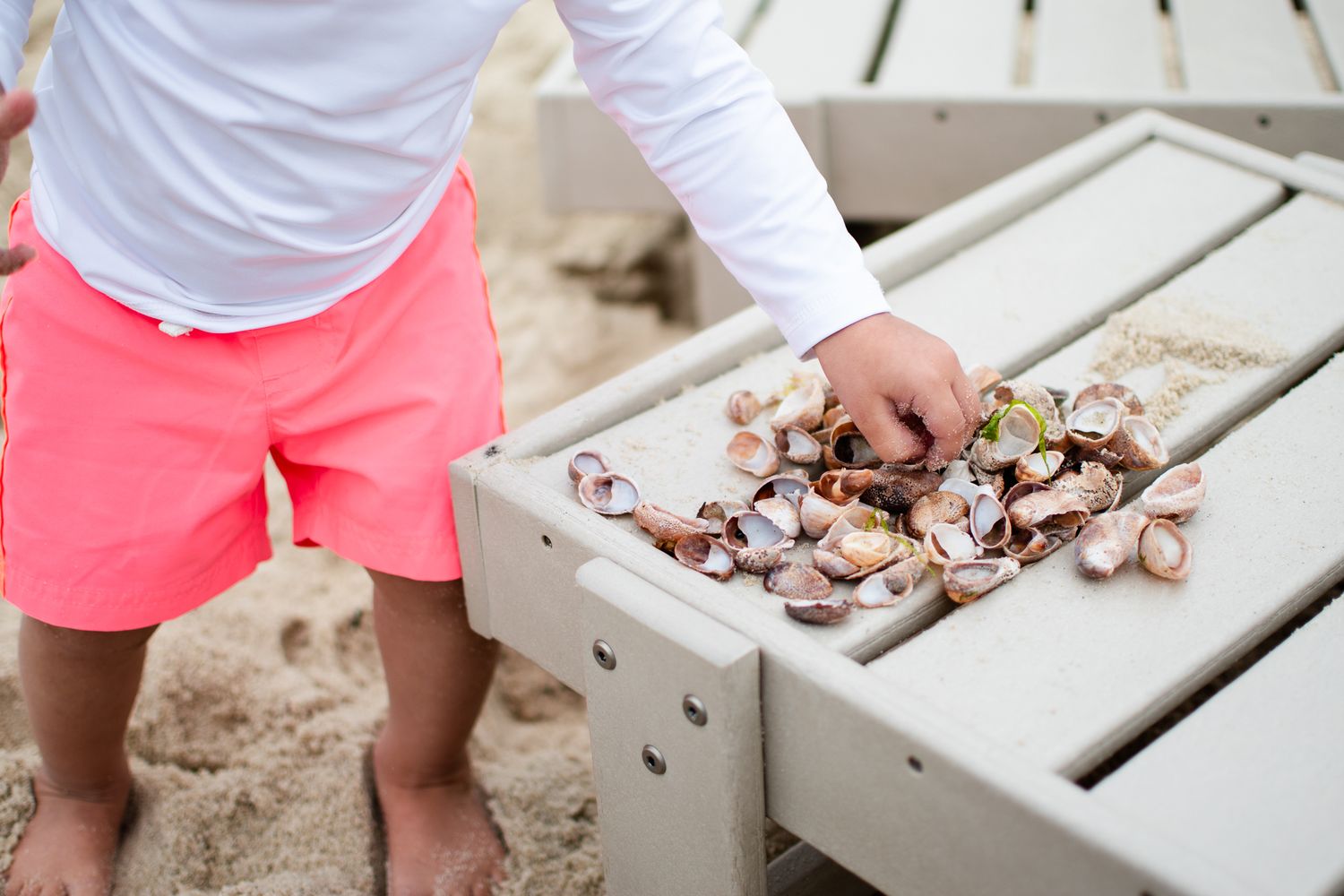Person wearing bright coral shorts sorts through fresh shellfish at a rustic outdoor table setup.