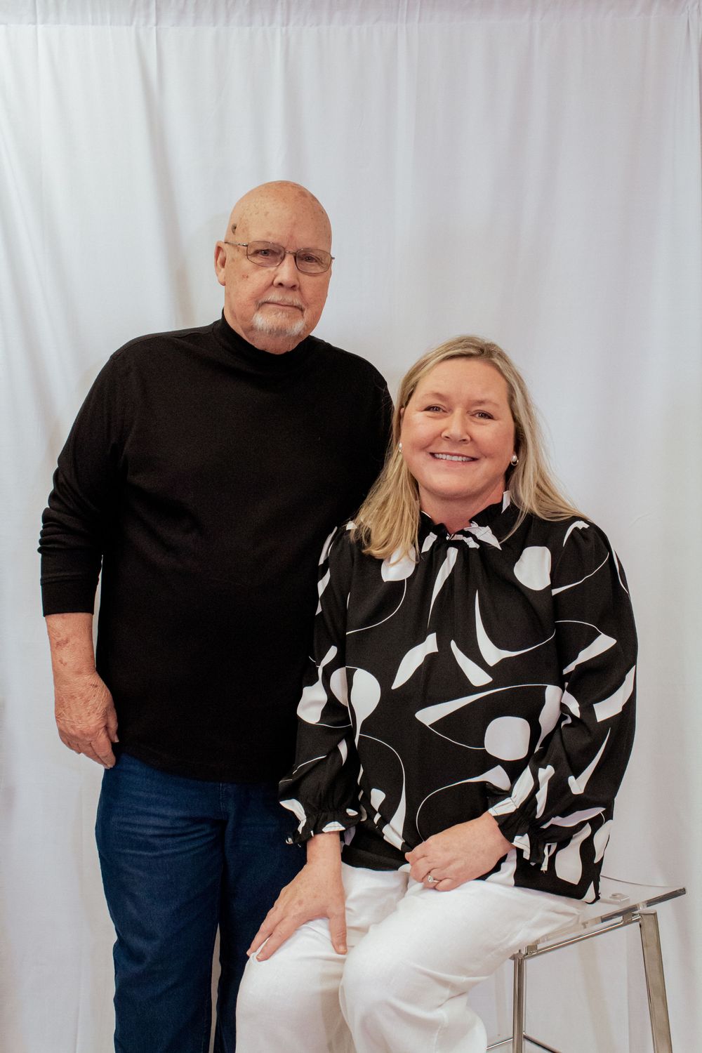 A couple poses in coordinating black and white outfits against a light background with a contemporary chair.