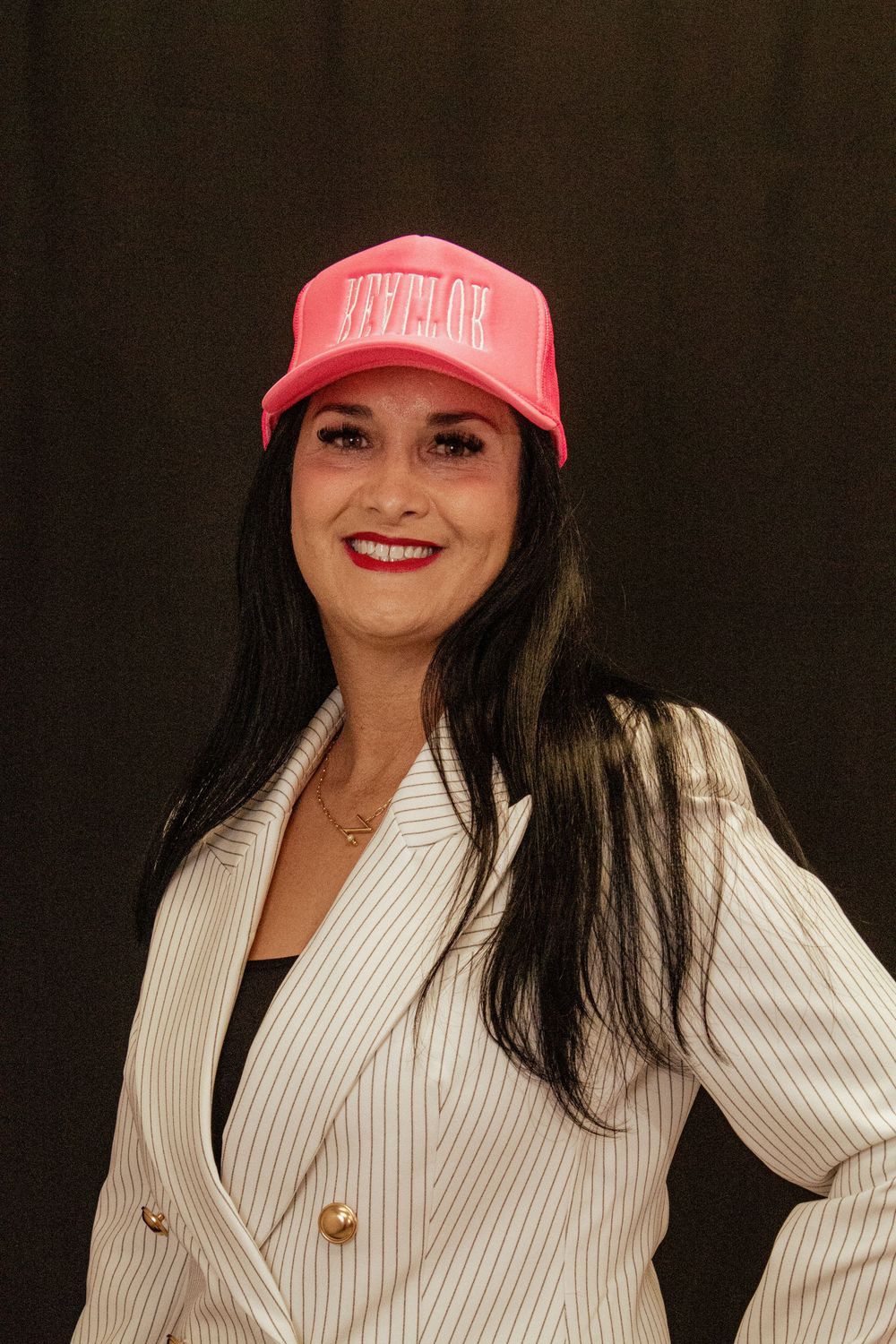 A person wearing a white pinstriped blazer and pink baseball cap smiles at the camera.