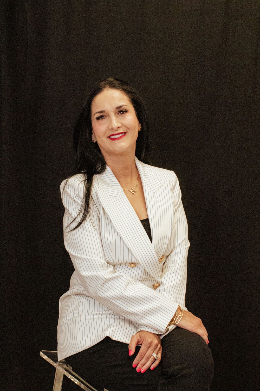 Professional portrait of someone in a crisp white pinstriped blazer and dark pants seated against a black backdrop.
