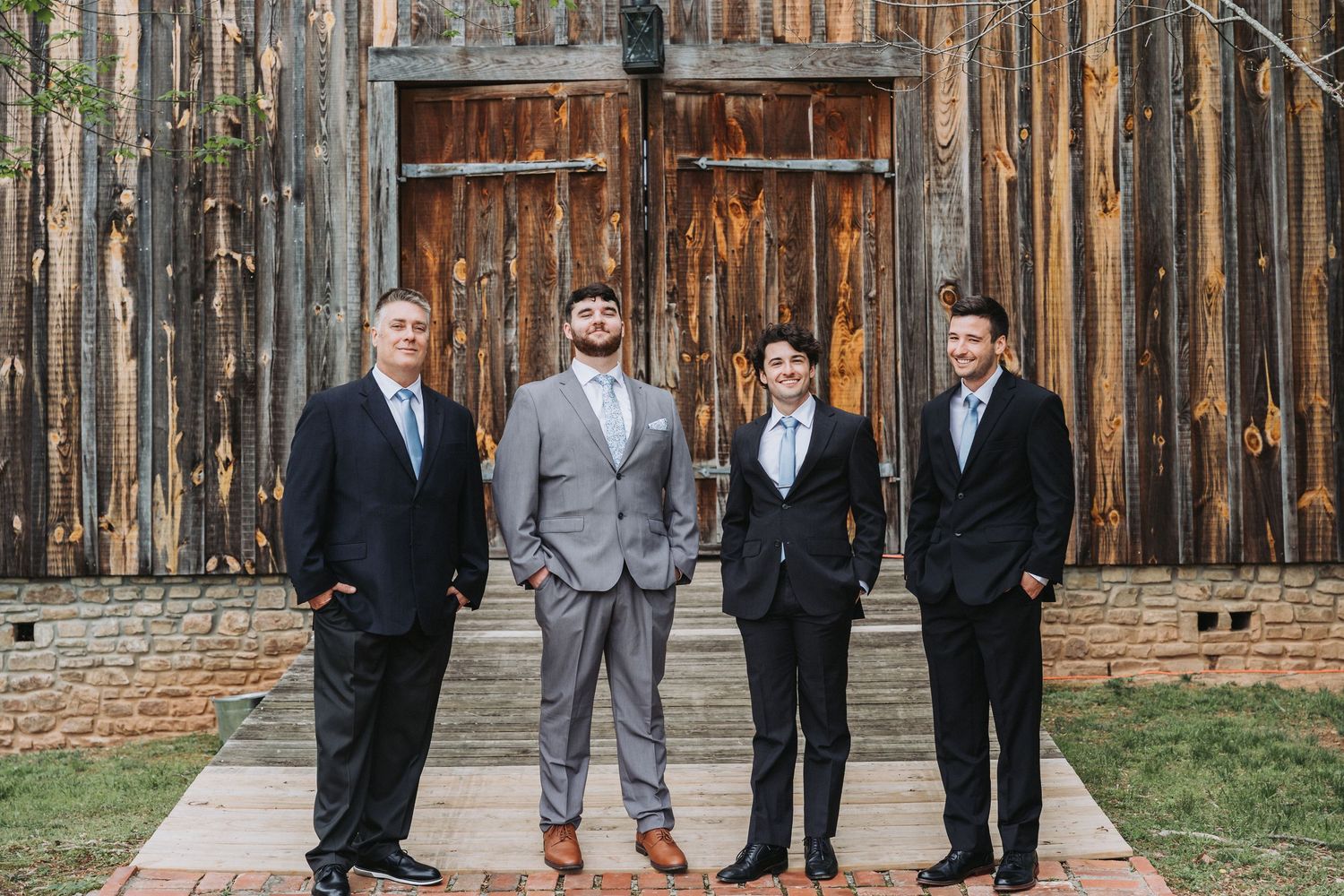 Four people in formal attire stand together in front of rustic wooden barn doors at a wedding venue.
