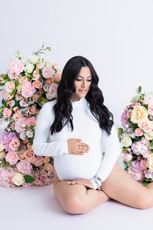Maternity photo featuring a white bodysuit against pink and peach floral arrangements on light background.