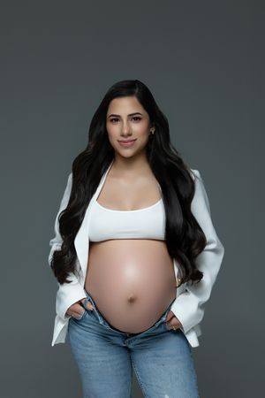 A maternity photo series showing a pregnant belly in white crop top and jeans against a dark gray backdrop.