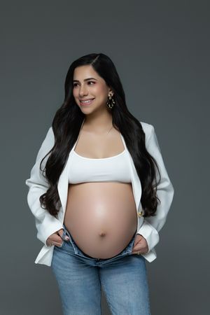Maternity photo of an expectant mother in white crop top and jeans with long dark hair against gray backdrop.
