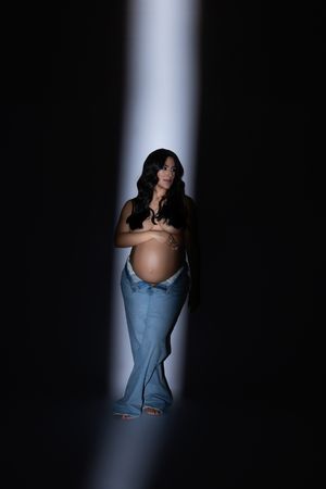 A pregnant figure poses in denim jeans against a dark background with dramatic spotlight illumination.