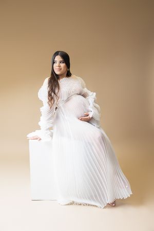 A person in a flowing white pleated dress with long sleeves poses elegantly against a beige backdrop.
