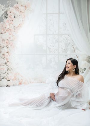Ethereal maternity photo series with flowing white fabric and delicate pink floral arrangements in a bright studio setting.