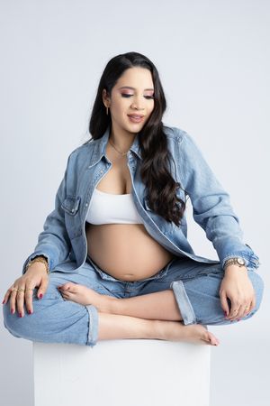 Woman in denim outfit sits cross-legged showcasing maternity style against white background.