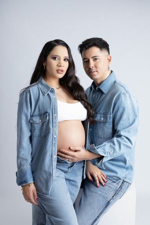Expectant couple in matching denim outfits pose together in studio setting.