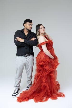 A man in a black shirt and gray pants stands with crossed arms beside a woman in a stunning red off-shoulder tulle gown.