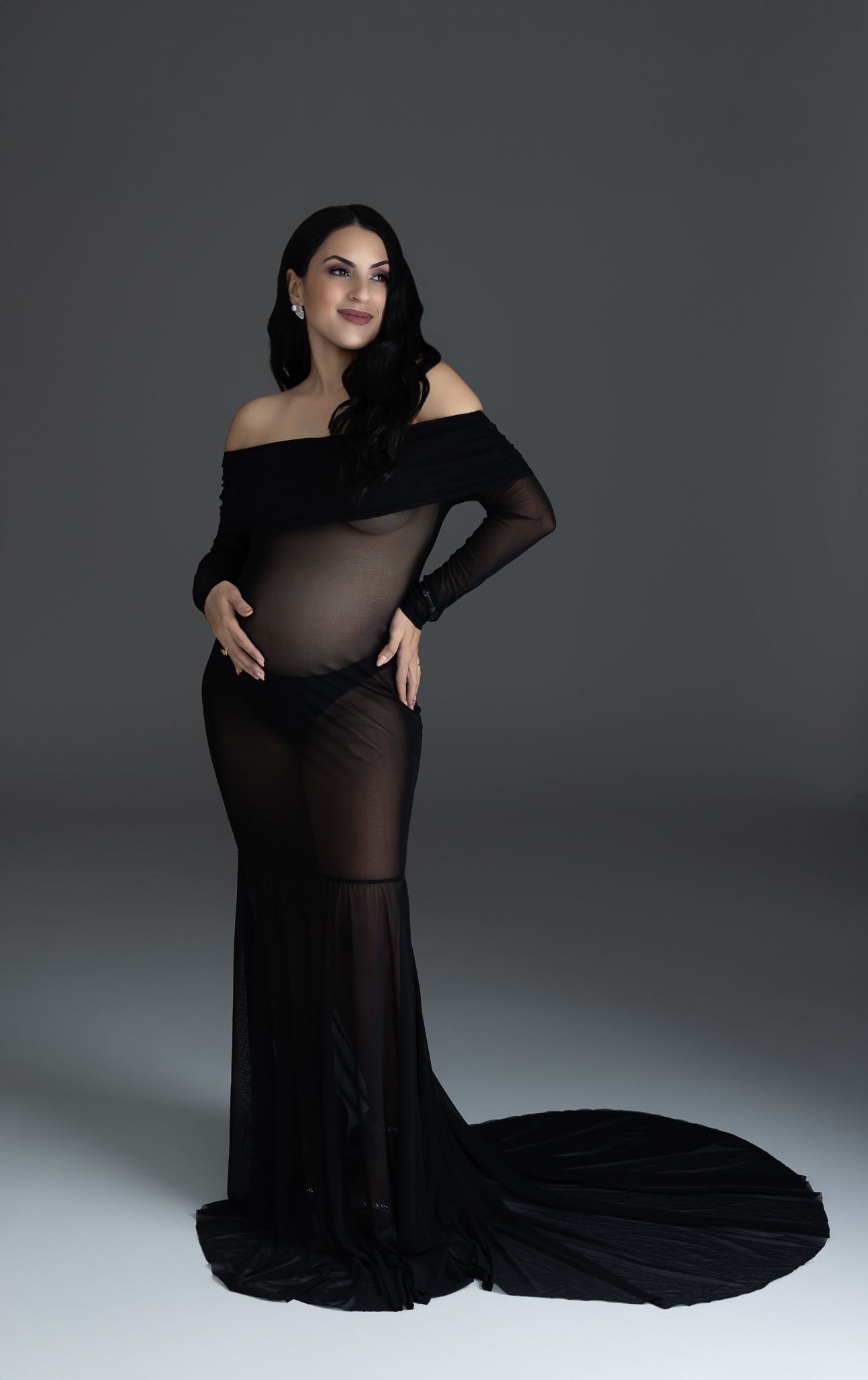 Pregnant woman in elegant black off-shoulder sheer maternity gown posing gracefully in a professional studio setting.