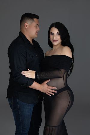 A pregnant couple in black outfits poses for a maternity photo shoot against a gray studio background.
