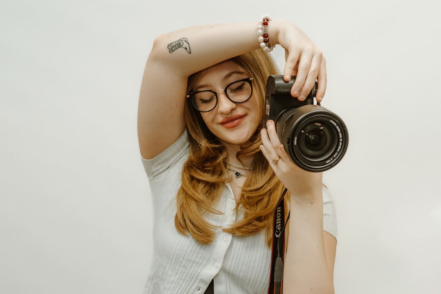 A photographer in round glasses and white sweater holds up a professional camera with a telephoto lens against a white background.
