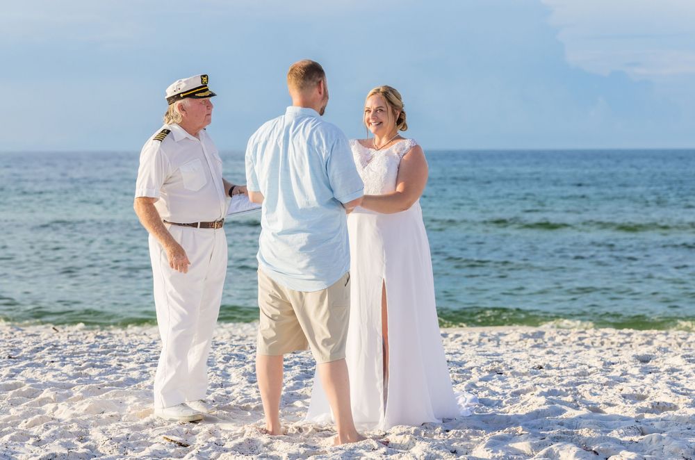 Three people in white clothing stand together on a sandy beach with the ocean and blue sky in the background.