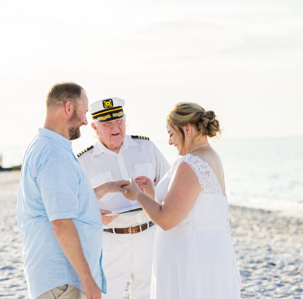 A marriage ceremony takes place on a sunny beach with waves in the background as a uniformed officiant conducts the service.