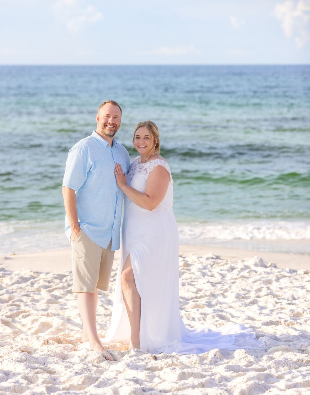 A couple poses on a scenic white sand beach with turquoise ocean waves in the background on a sunny day.