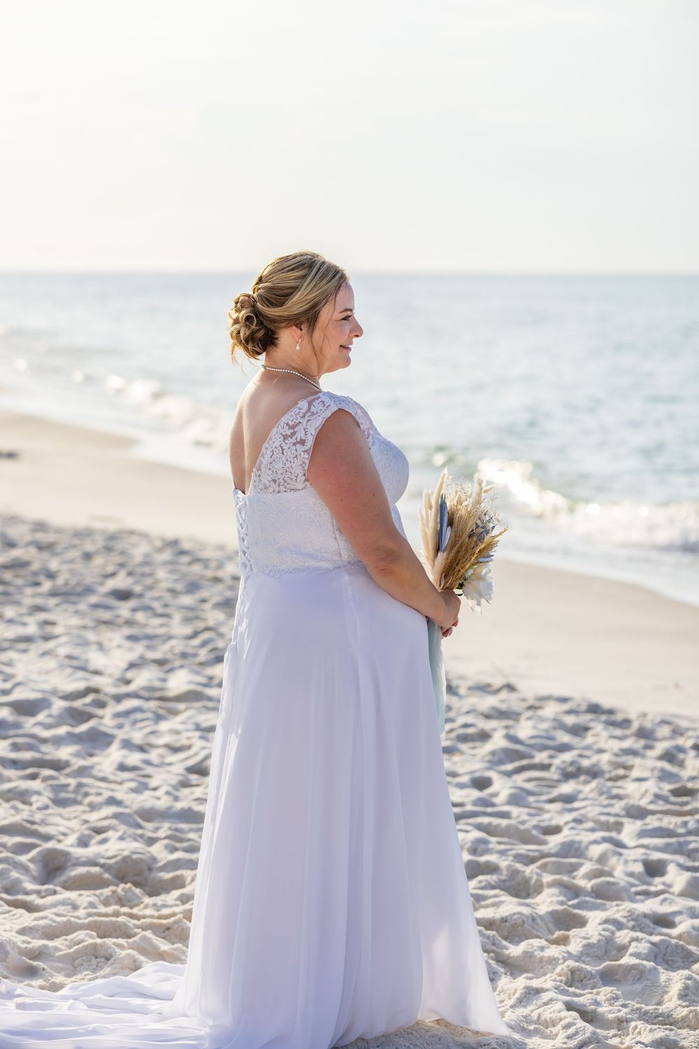 A bride in a white lace gown holds a dried flower bouquet while standing on a sunny beach at sunset.