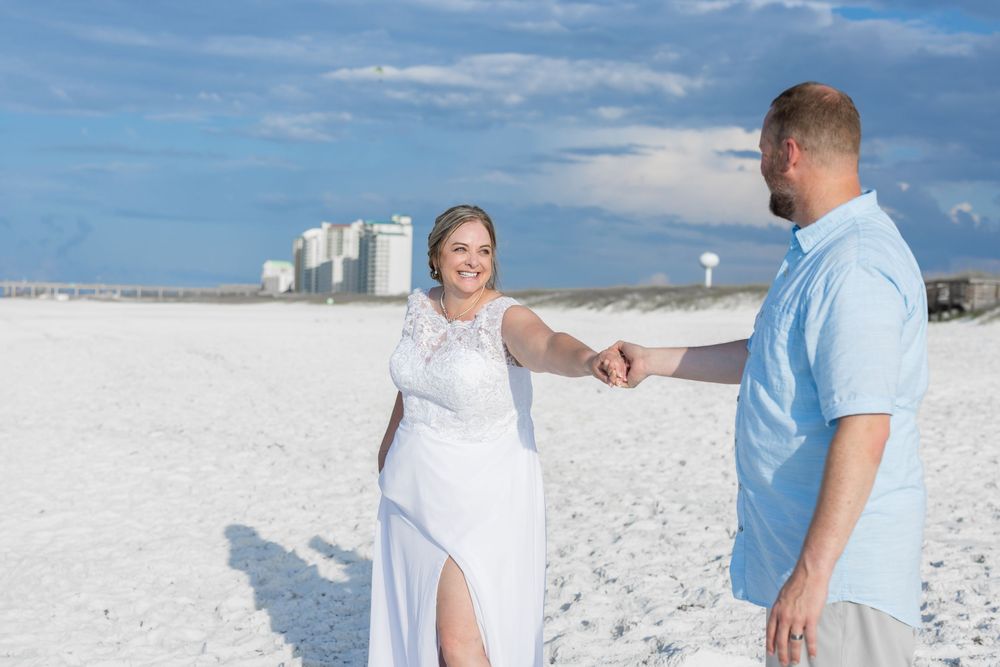 Two people hold hands on a bright white sandy beach with tall buildings visible in the background on a sunny day.