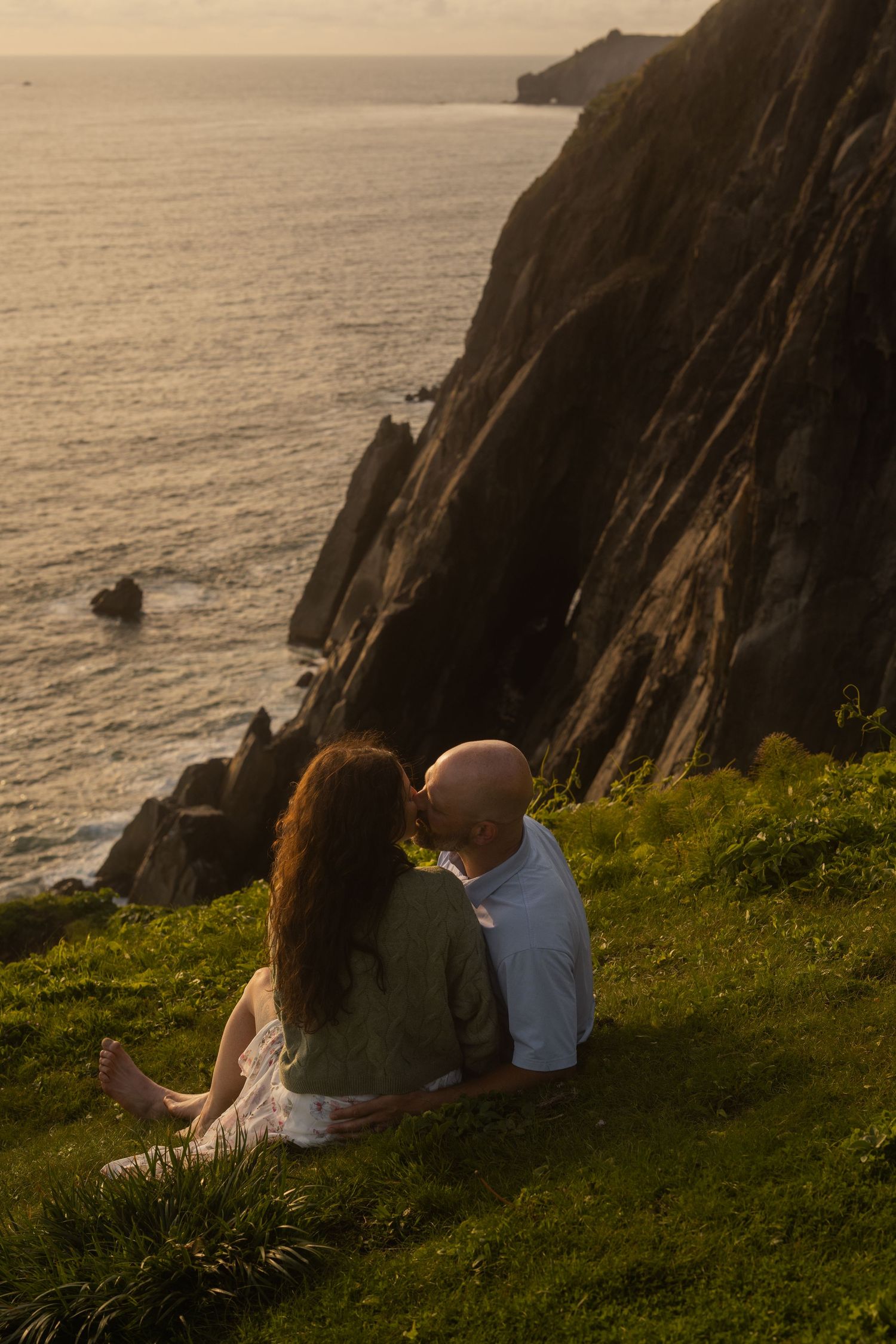 A couple sits on a grassy cliff overlooking dramatic coastal rocks and ocean at sunset in warm golden light.