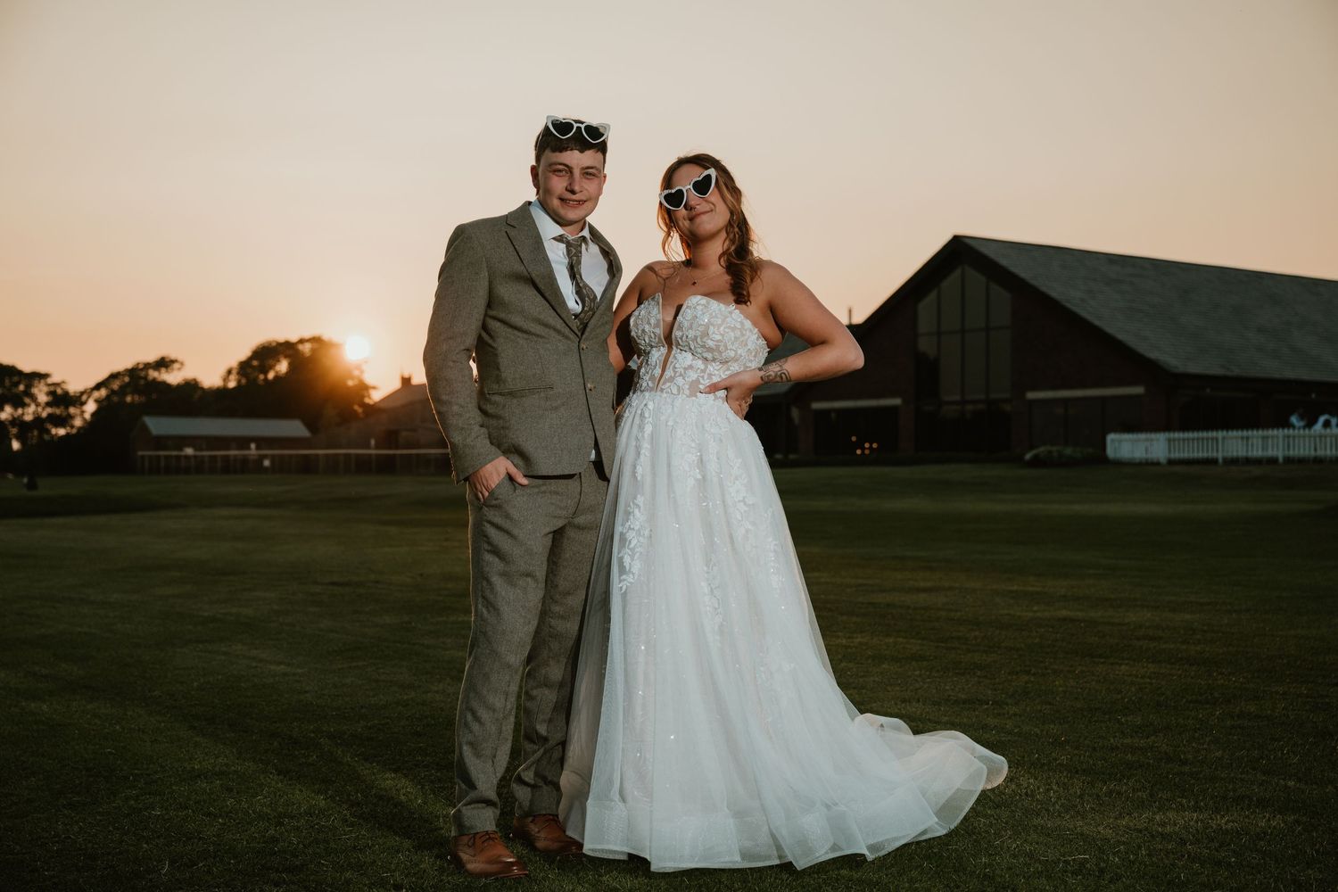 Wedding couple poses playfully in an open field at sunset with the backdrop of rural buildings.